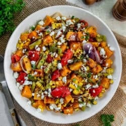 A bowl of roasted fall harvest salad with roasted butternut squash, corn, asparagus, red peppers, and red onion topped with crumbled feta and balsamic glaze.