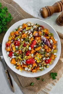 A bowl of roasted fall harvest salad with roasted butternut squash, corn, asparagus, red peppers, and red onion topped with crumbled feta and balsamic glaze.