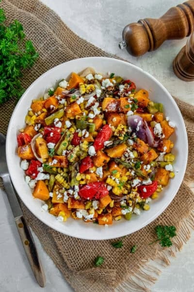 A bowl of roasted fall harvest salad with roasted butternut squash, corn, asparagus, red peppers, and red onion topped with crumbled feta and balsamic glaze.