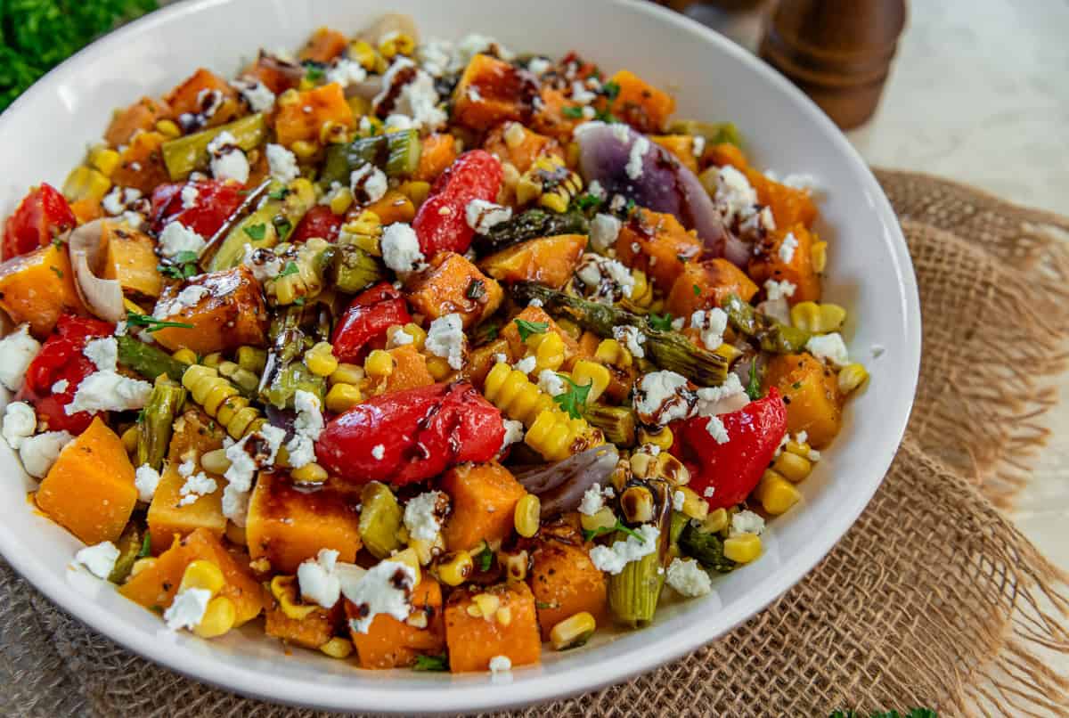 A bowl of roasted fall harvest salad with roasted butternut squash, asparagus, corn, red peppers, and red onion topped with crumbled feta and balsamic glaze.