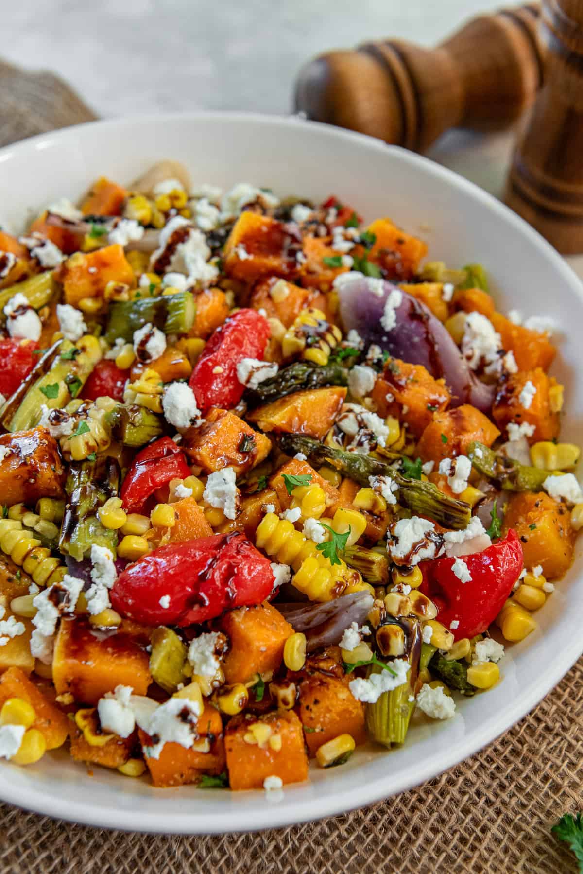 A large bowl of warm roasted vegetables including butternut squash, corn, asparagus, red peppers, and red onion topped with crumbled goat cheese and balsamic glaze.