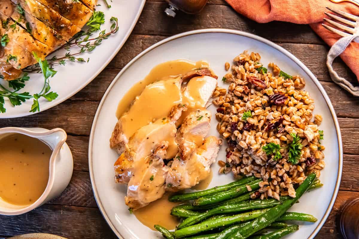 Sliced maple glazed turkey breast served with pan gravy, green beans, and farro pilaf with toasted nuts on a dinner plate.