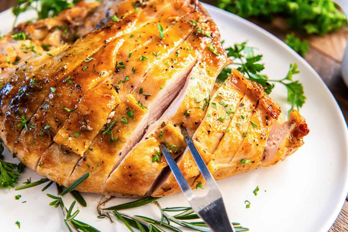 Maple glazed turkey breast sliced on a serving platter with rosemary and parsley, showing crisp skin and tender meat.