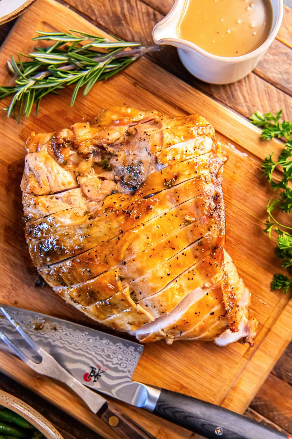 Maple glazed turkey breast sliced on a wooden cutting board with fresh herbs, a carving knife, and gravy nearby.