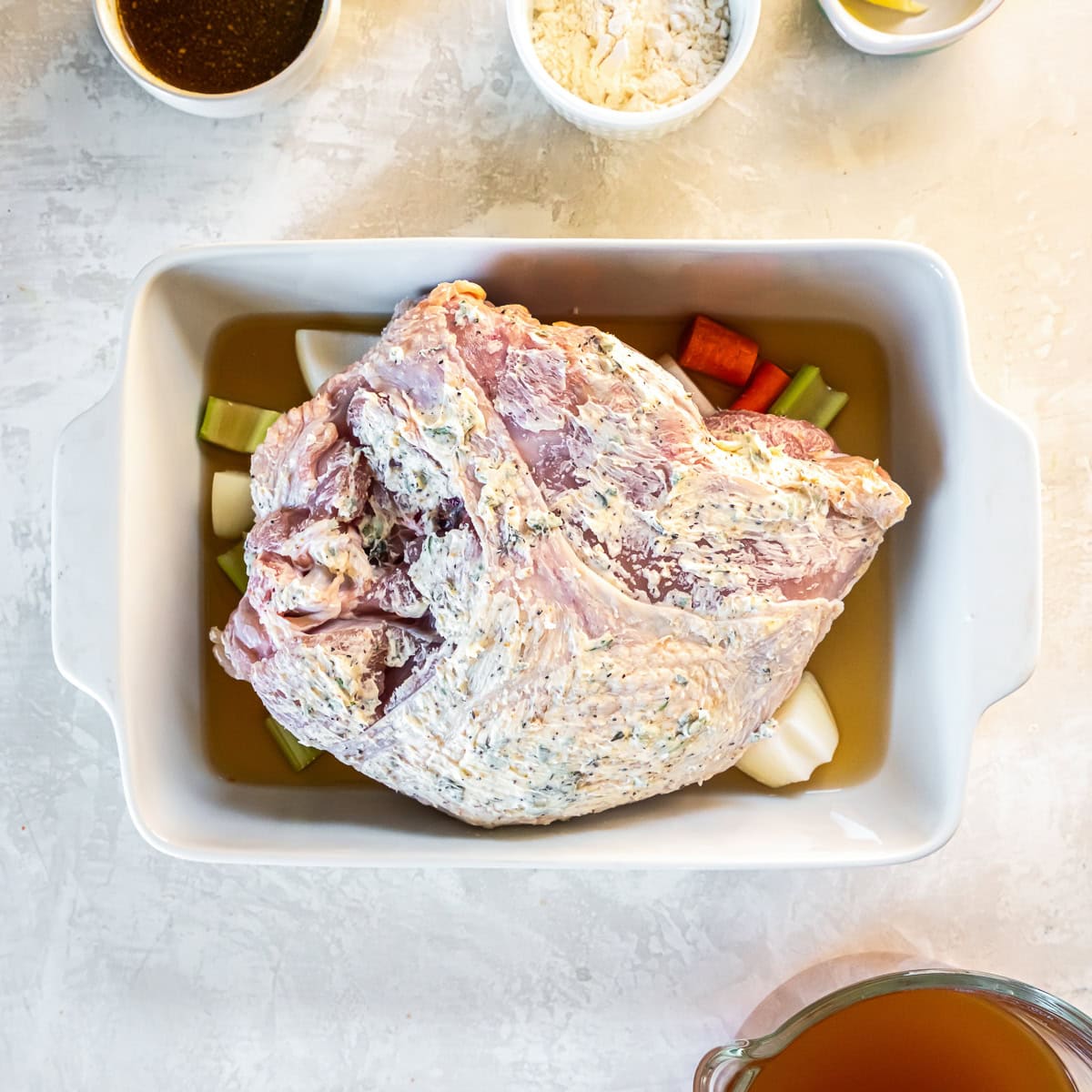 Raw bone-in turkey breast coated with herb butter and placed on vegetables in a baking dish with broth.
