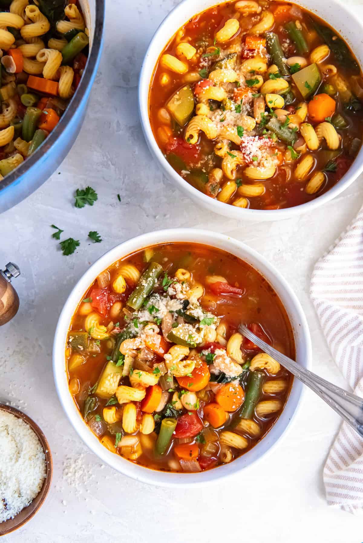 Two bowls of vegetable white bean protein pasta soup topped with Parmesan and fresh parsley, with colorful vegetables and curly protein pasta in a rich tomato broth.