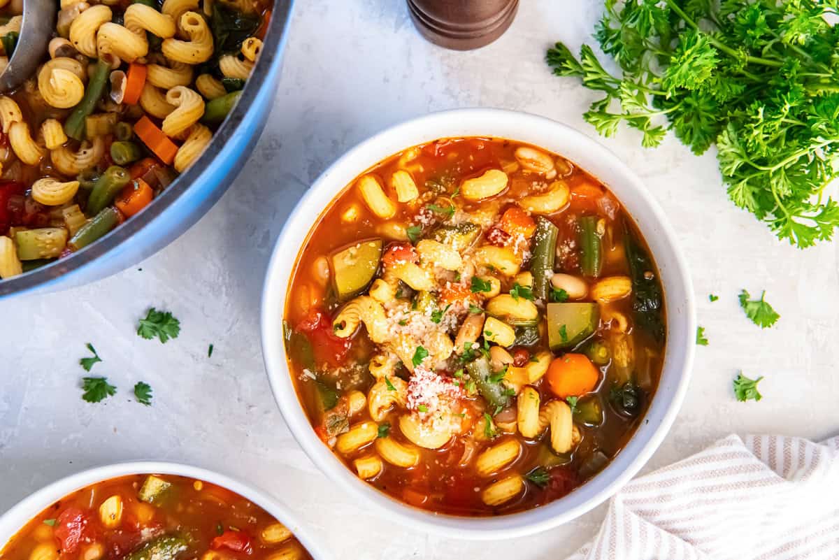 Bowl of vegetable protein pasta soup topped with grated Parmesan and fresh parsley, with cannellini beans, curly mac protein pasta, carrots, zucchini, green beans, and tomatoes in a tomato broth.