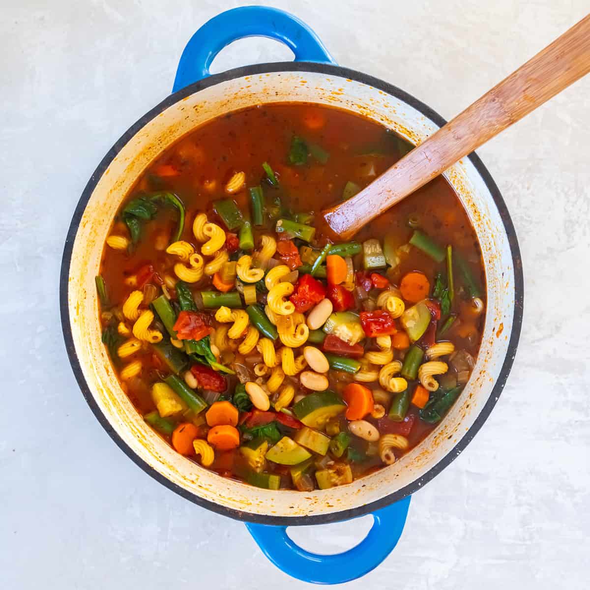 Vegetable white bean protein pasta soup simmering in a Dutch oven with curly mac pasta, cannellini beans, zucchini, carrots, green beans, tomatoes, and spinach in a tomato broth.