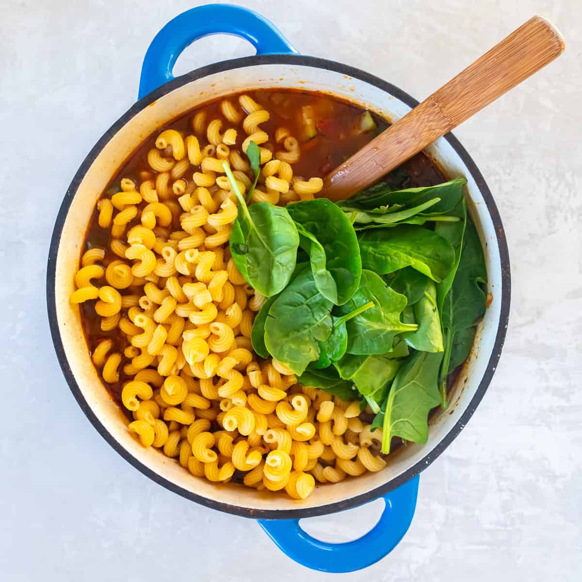 Cooked protein pasta and fresh baby spinach added to a pot of vegetable soup before stirring