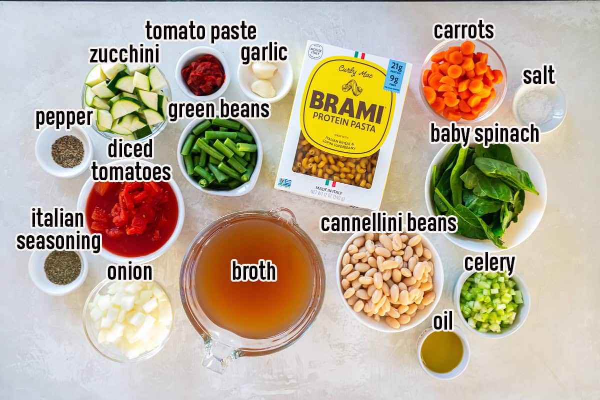 Ingredients for vegetable white bean protein pasta soup arranged on a light surface, including protein pasta, cannellini beans, carrots, celery, onion, zucchini, green beans, spinach, garlic, diced tomatoes, tomato paste, olive oil, broth, and seasonings.