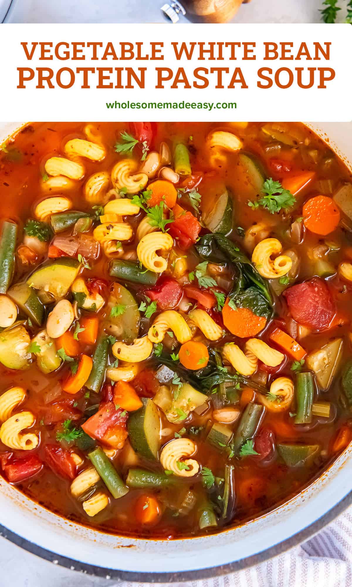 Vegetable protein pasta soup in a Dutch oven with cannellini beans, curly mac protein pasta, carrots, zucchini, green beans, tomatoes, and spinach in a tomato broth, garnished with fresh parsley with text overlay.
