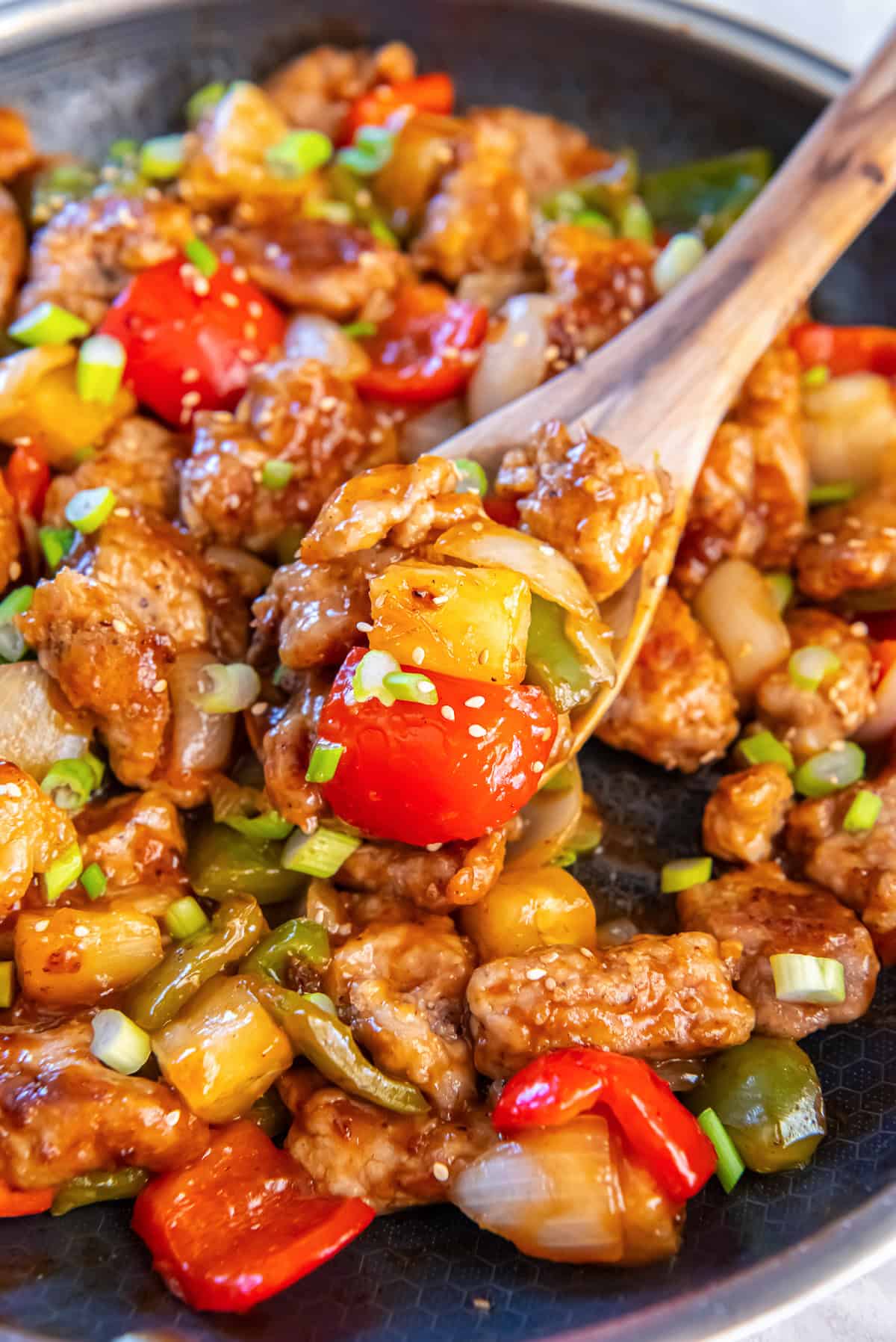 A wooden spoon scoopiing sweet and sour pork in a skillet with bell peppers and pineapple.