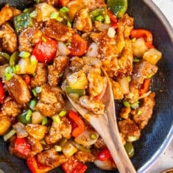 Sweet and sour pork in a skillet with bell peppers and pineapple, shown with a wooden spoon.
