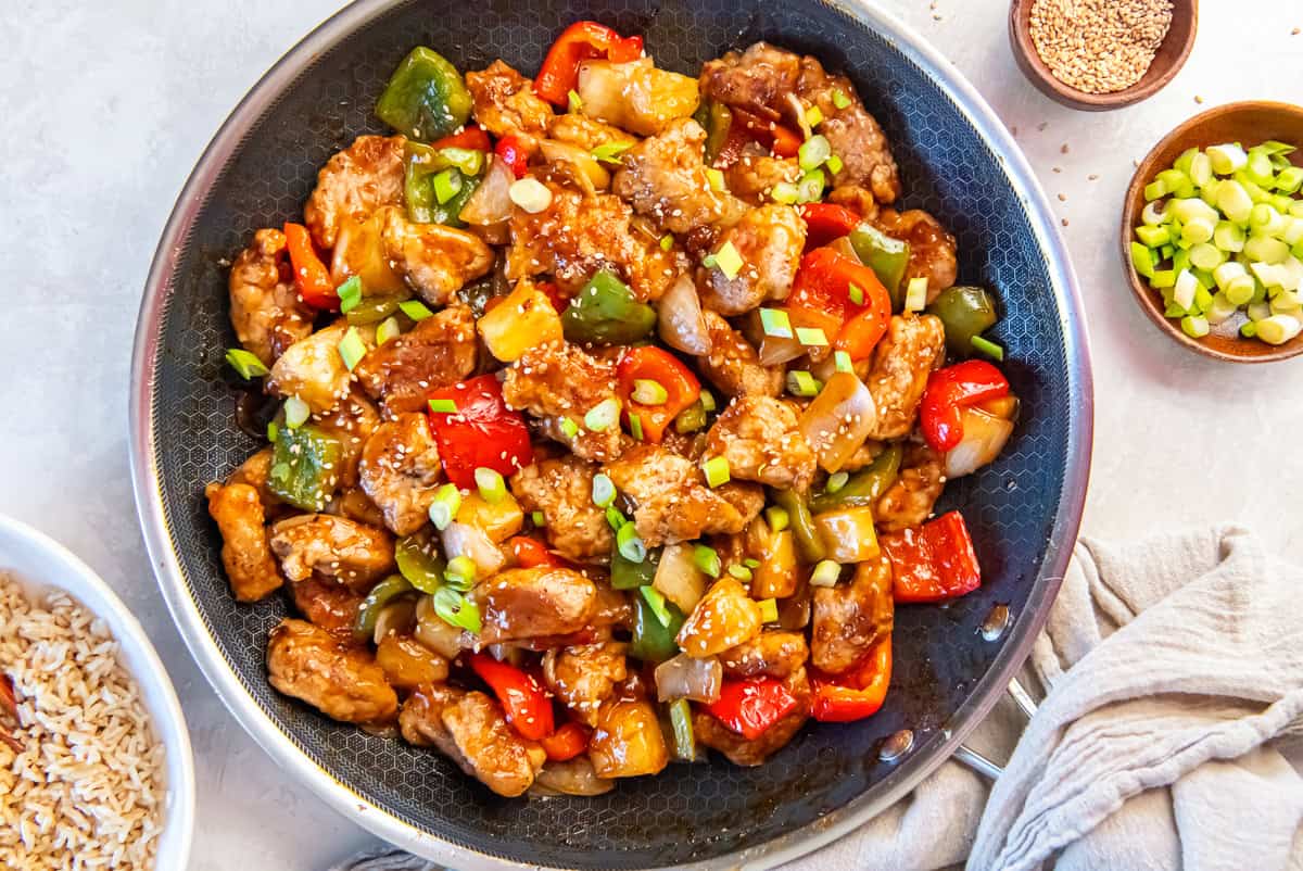 sweet and sour pork with bell peppers and pineapple in a skillet, served with brown rice and garnished with green onions.