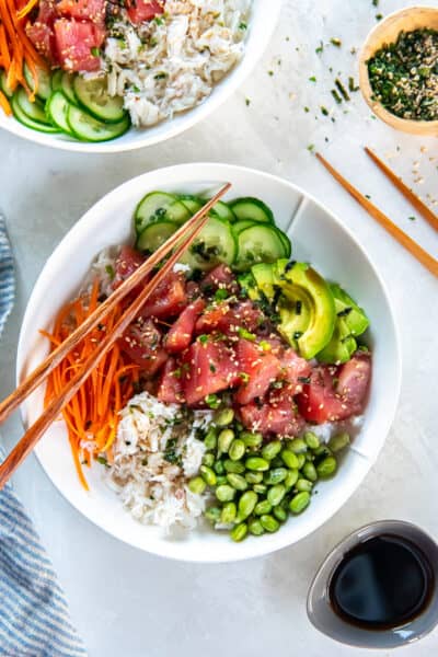 Ahi tuna poke bowl with marinated tuna, avocado, cucumber, carrot, edamame, and crab over white rice with chopsticks.
