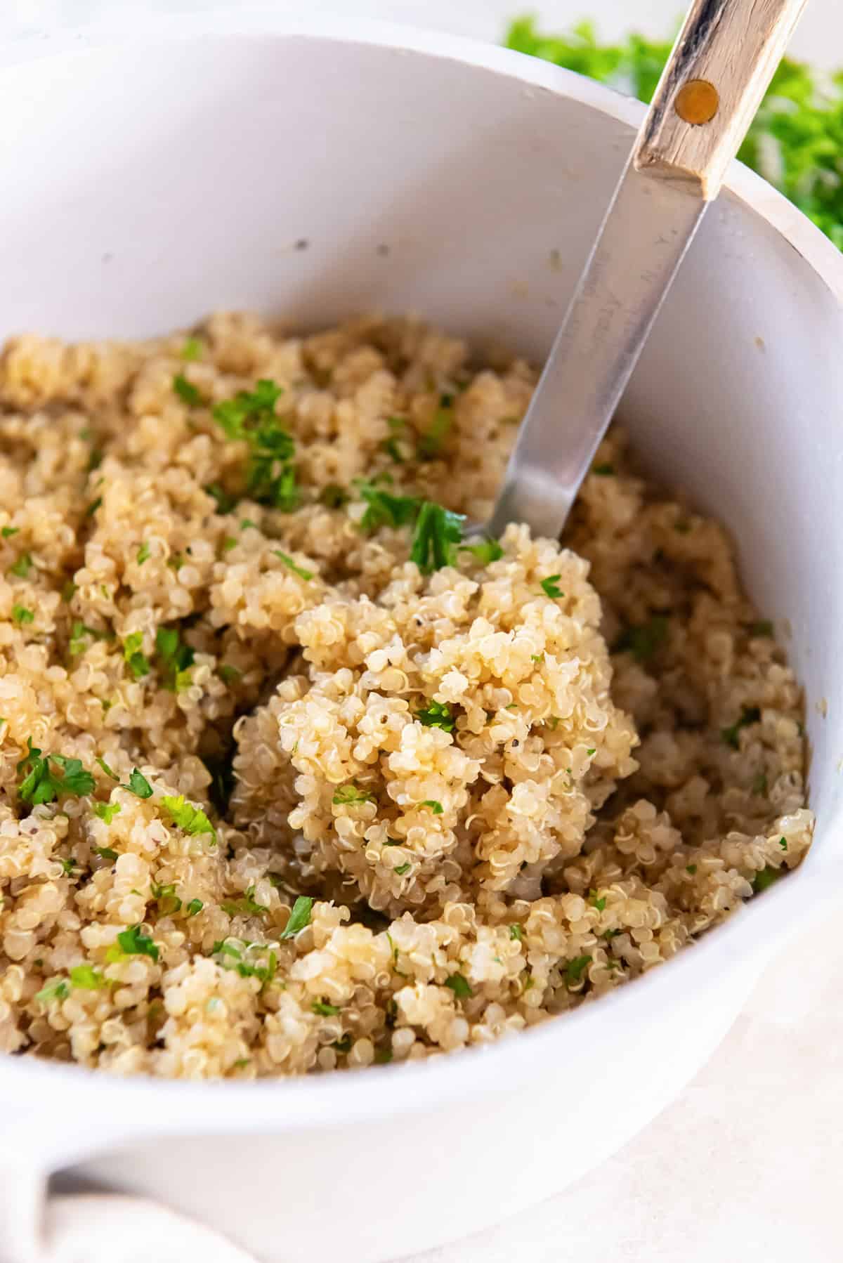 Spoon scooping fluffy garlic butter quinoa from a white saucepan with parsley garnish.