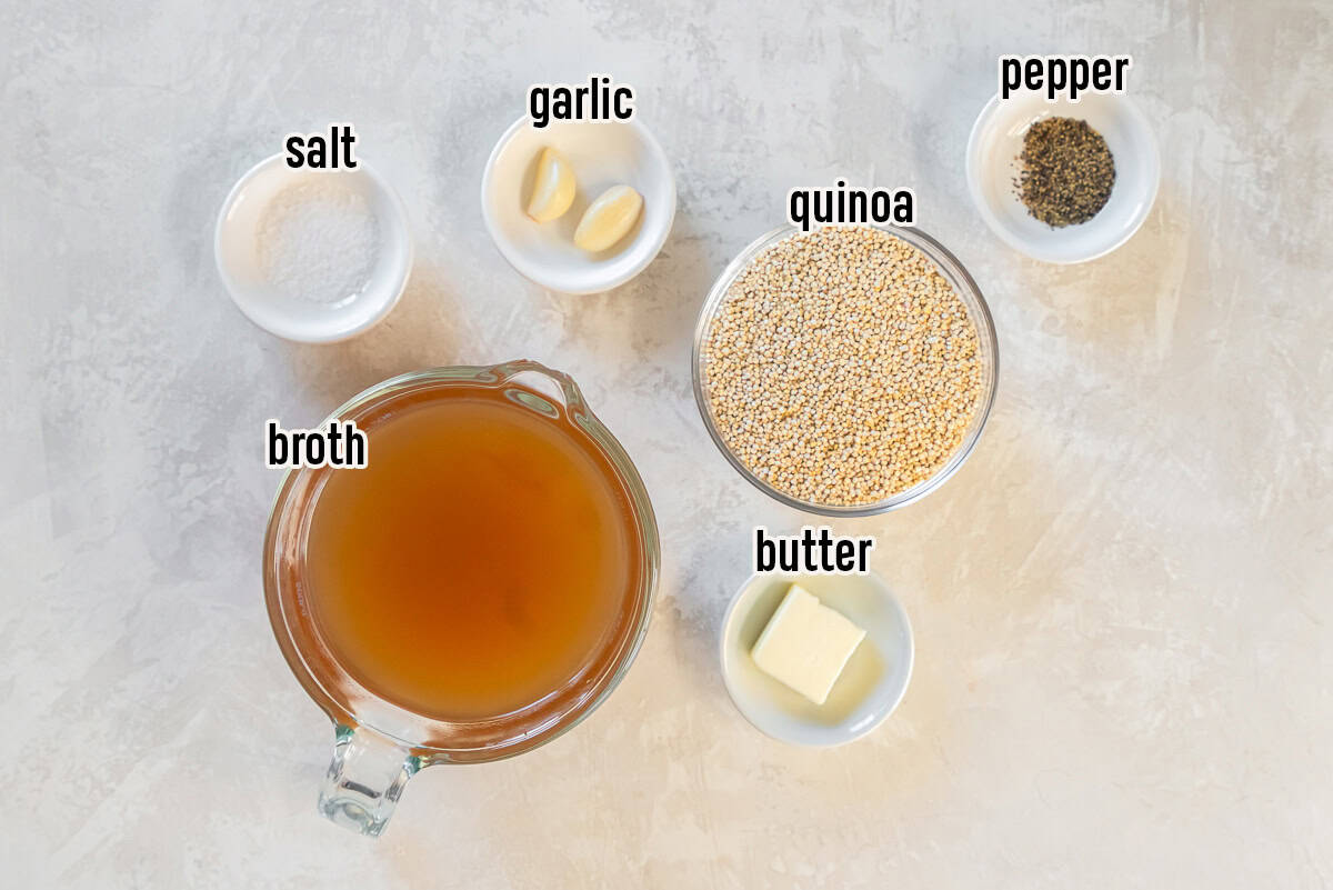 Ingredients for garlic butter quinoa including quinoa, broth, butter, garlic cloves, salt, and black pepper in small bowls on a light surface.