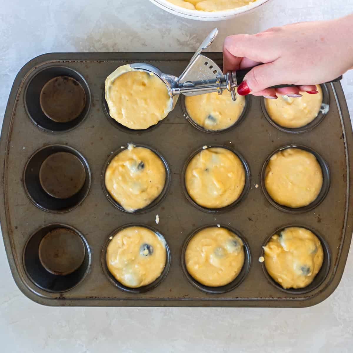 Scooping blueberry muffin batter into a greased muffin pan.