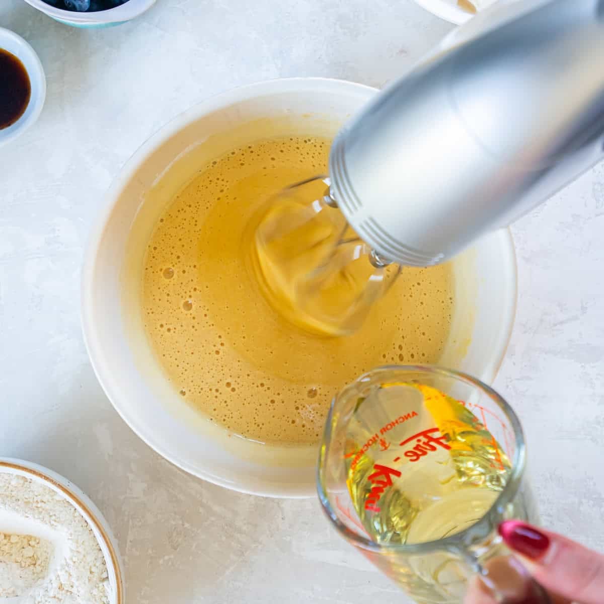 Beating eggs and sugar with an electric mixer while adding oil for blueberry muffin batter.