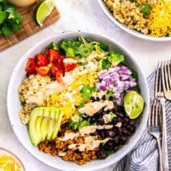 two Chicken Taco Grain Bowls topped with seasoned ground chicken, quinoa, black beans, avocado, cherry tomatoes, shredded cheese, red onion, romaine lettuce, and creamy cilantro lime dressing.