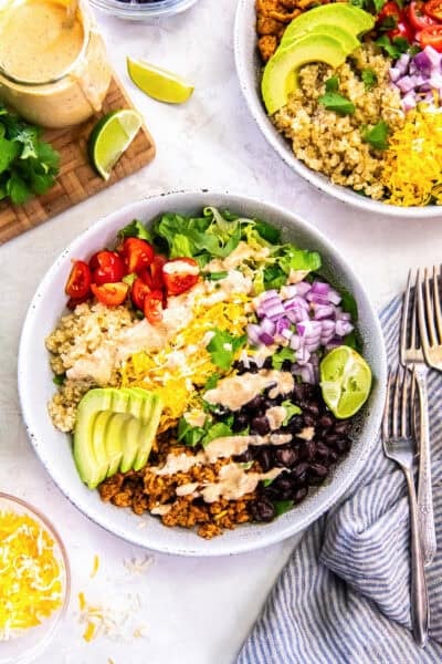 two Chicken Taco Grain Bowls topped with seasoned ground chicken, quinoa, black beans, avocado, cherry tomatoes, shredded cheese, red onion, romaine lettuce, and creamy cilantro lime dressing.