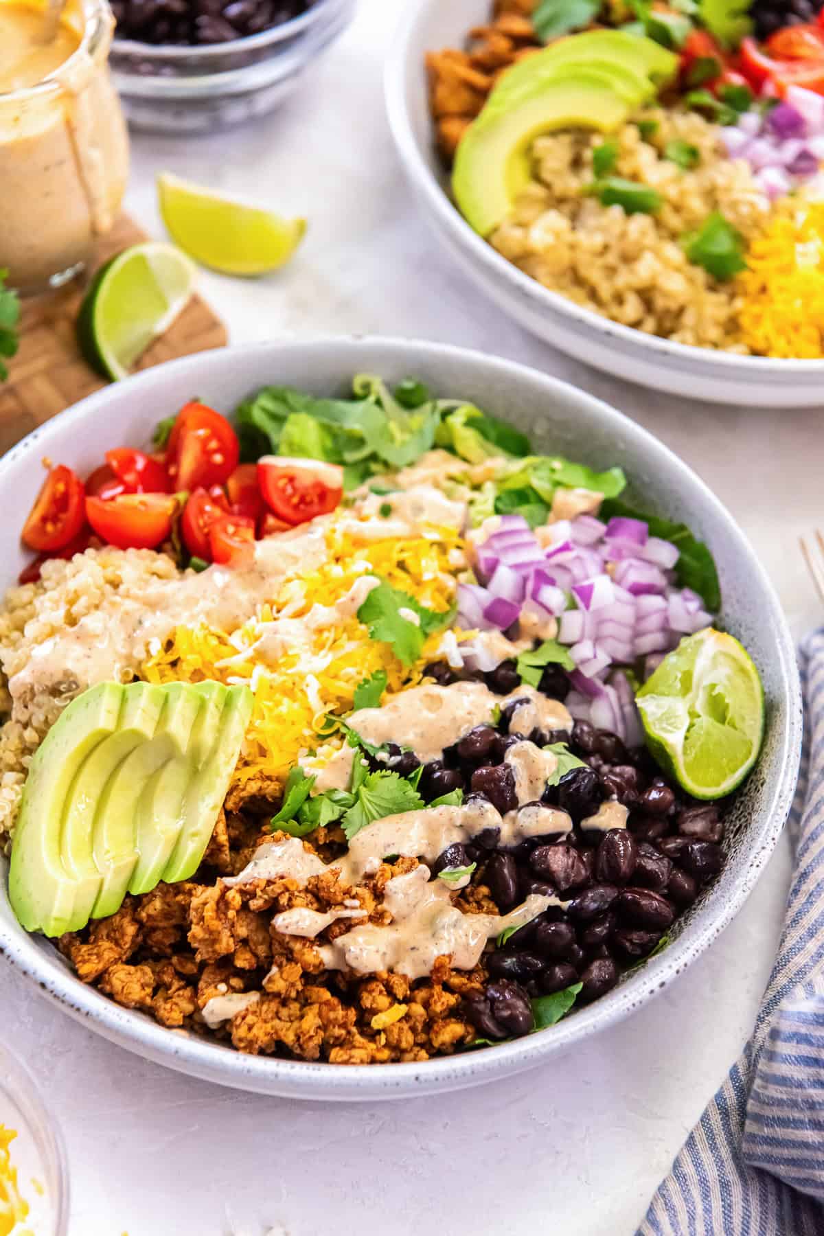 A Chicken Taco Grain Bowl with seasoned ground chicken, quinoa, black beans, avocado, cherry tomatoes, shredded cheese, red onion, romaine lettuce, lime wedges, and creamy cilantro lime dressing.