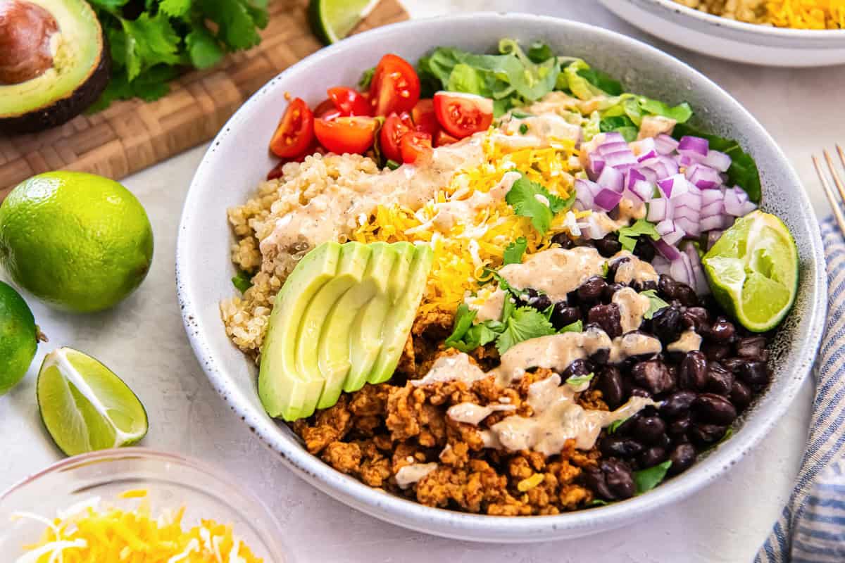 Chicken Taco Grain Bowls topped with seasoned chicken, black beans, quinoa, avocado, cheddar cheese, cherry tomatoes, red onion, and creamy taco dressing in a gray bowl with lime wedges and fresh cilantro.