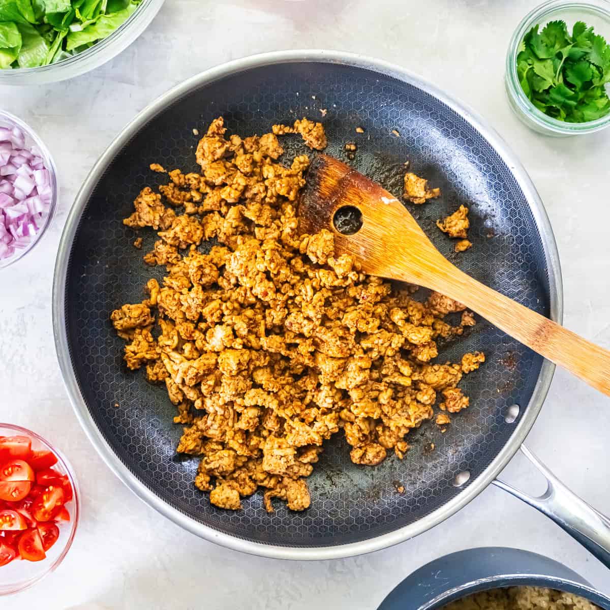 Seasoned ground chicken cooking in a skillet for Chicken Taco Grain Bowls with fresh bowl ingredients surrounding the pan.