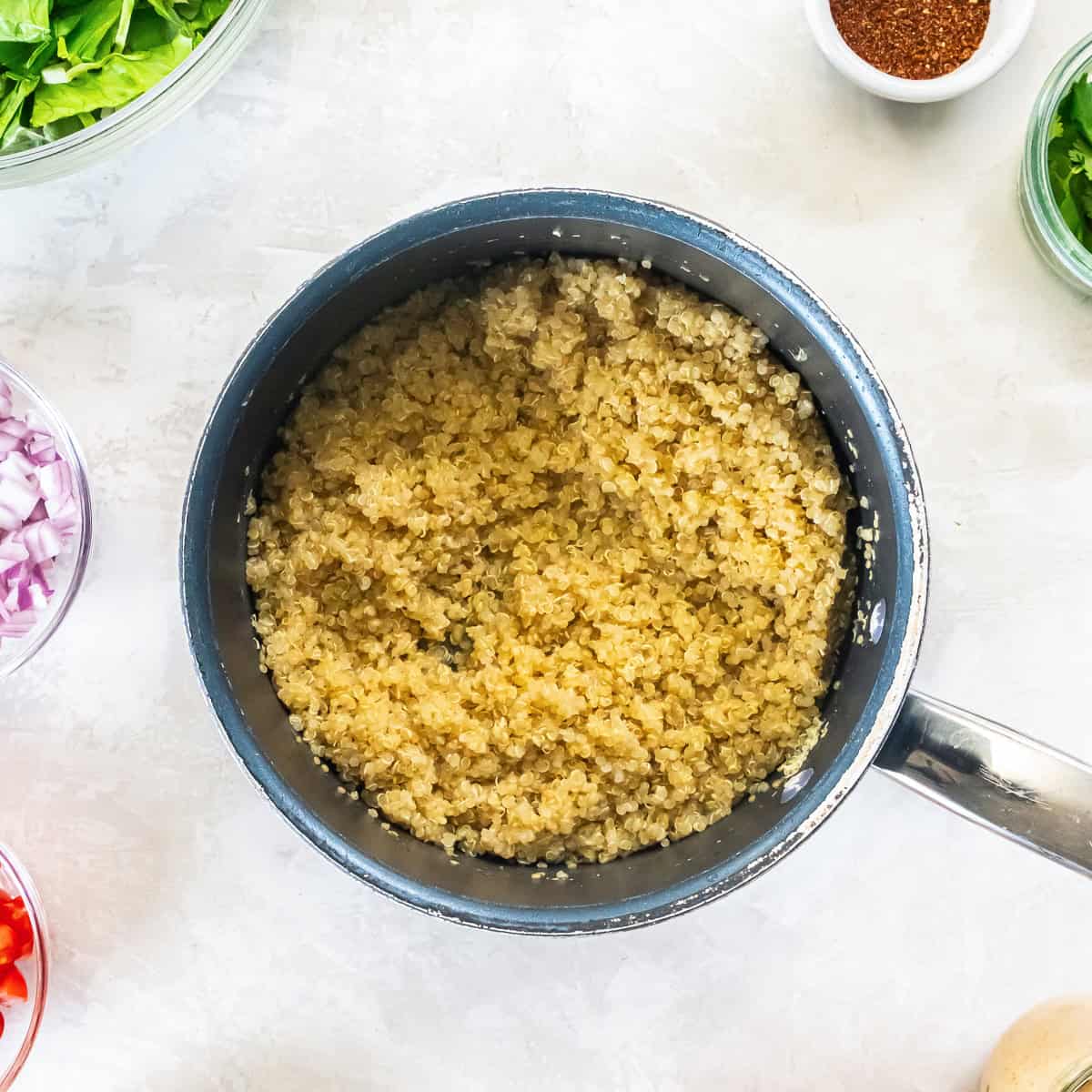 Cooked quinoa in a saucepan for Chicken Taco Grain Bowls with fresh taco bowl ingredients in the background.