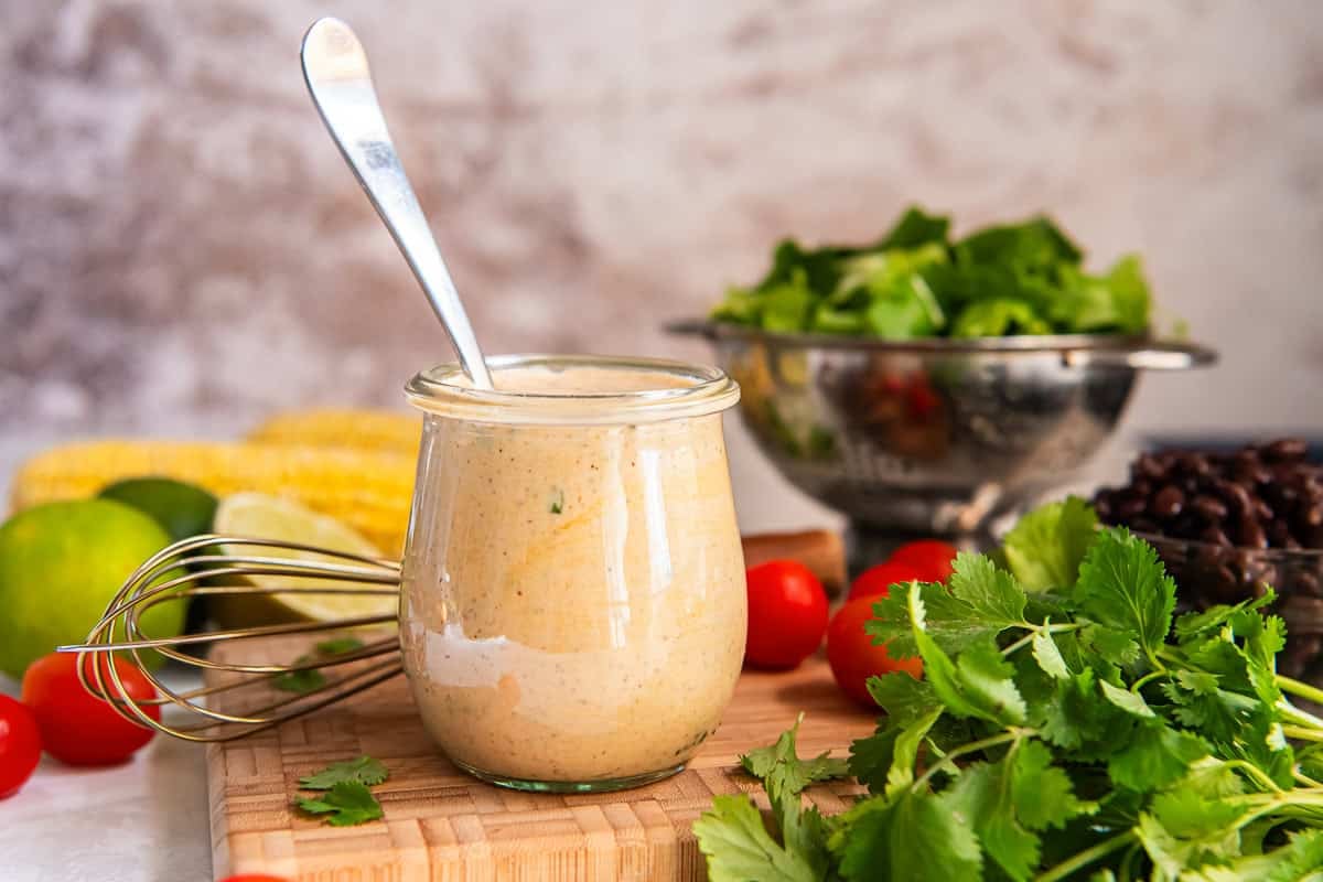 Glass jar of Creamy Cilantro Lime Dressing on a wooden board surrounded by fresh cilantro, tomatoes, limes, corn, lettuce, and black beans.