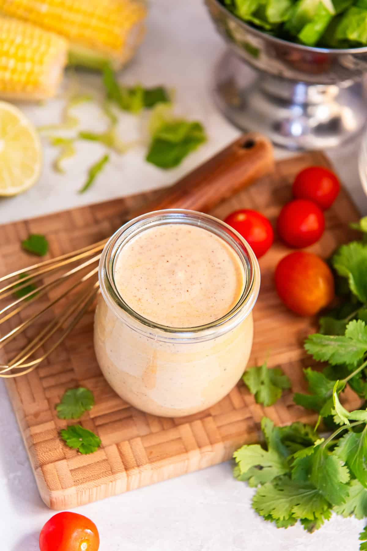 Glass jar of Creamy Cilantro Lime Dressing on a wooden board with fresh cilantro, cherry tomatoes, corn, and lime in the background.