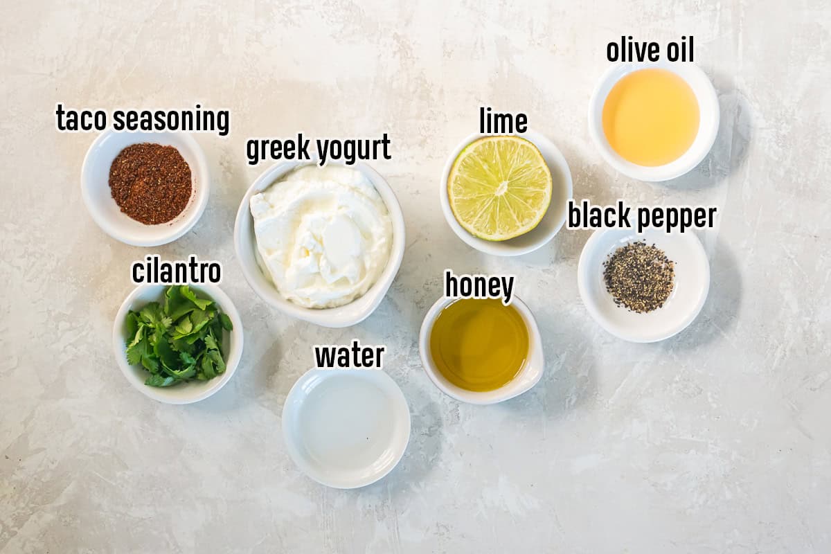 Ingredients for creamy cilantro lime dressing including Greek yogurt, cilantro, lime, olive oil, honey, taco seasoning, black pepper, and water in small bowls.