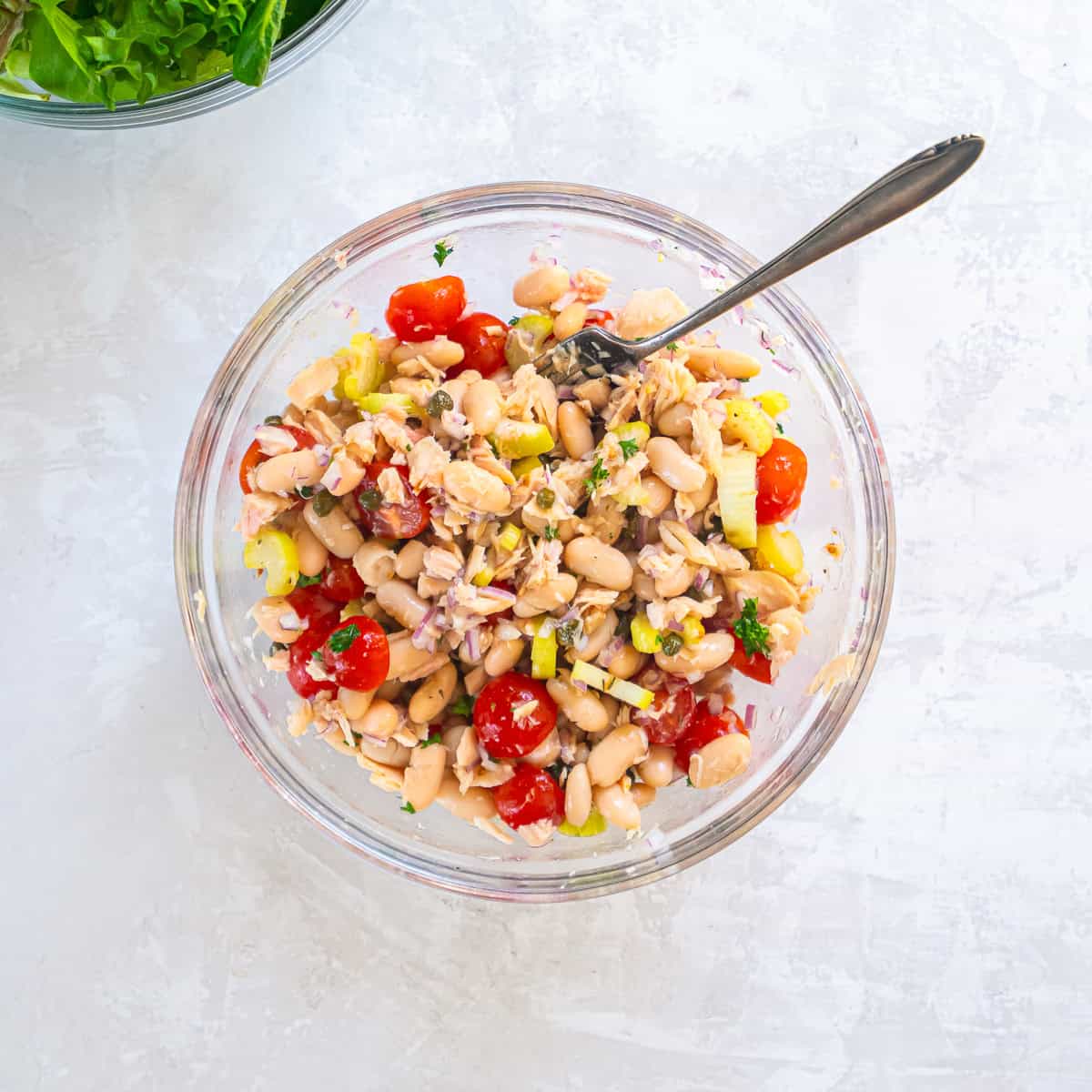 Tuna and white bean salad mixed in a glass bowl with cannellini beans, albacore tuna, cherry tomatoes, celery, red onion, capers, parsley, and vinaigrette.