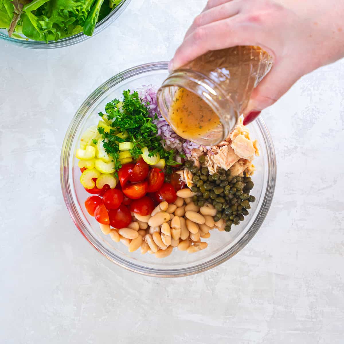 Red wine vinaigrette being poured over tuna, cannellini beans, cherry tomatoes, celery, red onion, capers, and parsley in a glass mixing bowl.