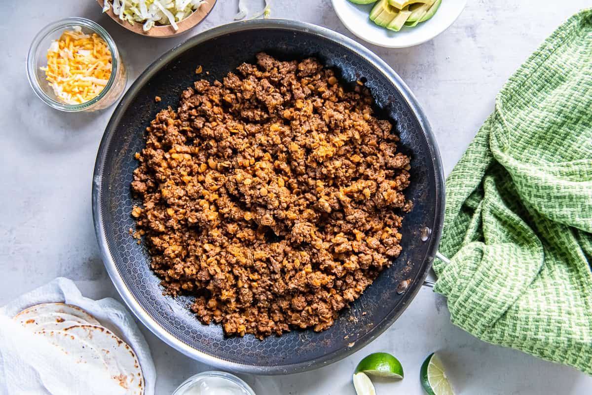 Ground beef taco meat with red lentils cooked in a large skillet with tortillas and taco toppings on the side.