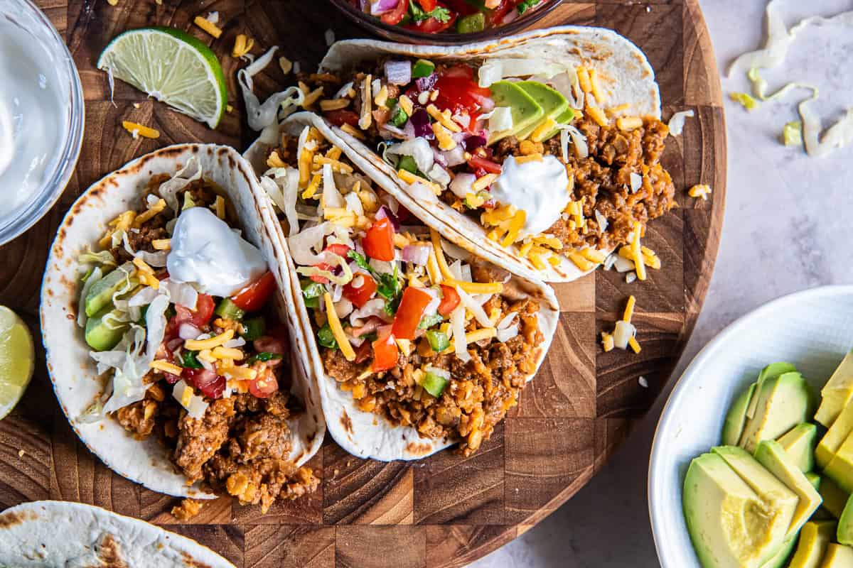 "Soft tacos filled with ground beef taco meat and red lentils, topped with cheese, pico de gallo, avocado, lettuce, and sour cream.