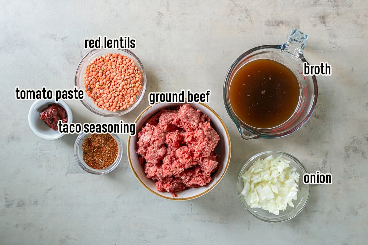 Ingredients for Ground Beef Taco Meat with Lentils arranged on a light surface, including lean ground beef, diced onion, beef broth, red split lentils, tomato paste, and taco seasoning.