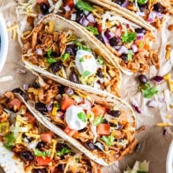 A row of Rotisserie Chicken Tacos topped with black beans, tomatoes, lettuce, cheese, red onion, cilantro, and sour cream on parchment paper.