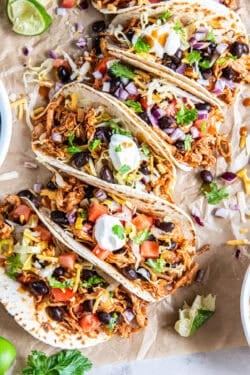 A row of Rotisserie Chicken Tacos topped with black beans, tomatoes, lettuce, cheese, red onion, cilantro, and sour cream on parchment paper.