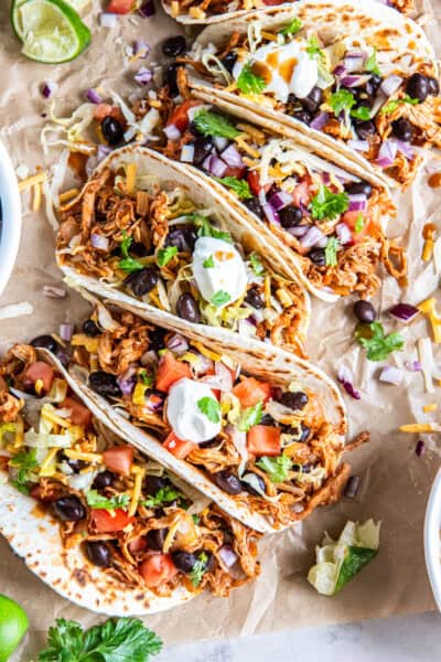 A row of Rotisserie Chicken Tacos topped with black beans, tomatoes, lettuce, cheese, red onion, cilantro, and sour cream on parchment paper.