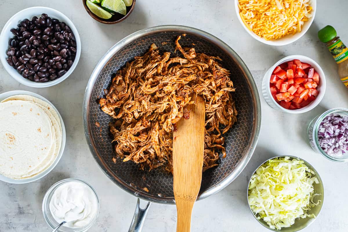 Seasoned shredded rotisserie chicken for tacos in a skillet surrounded by tortillas, black beans, lettuce, cheese, tomatoes, red onion, sour cream, and lime wedges.
