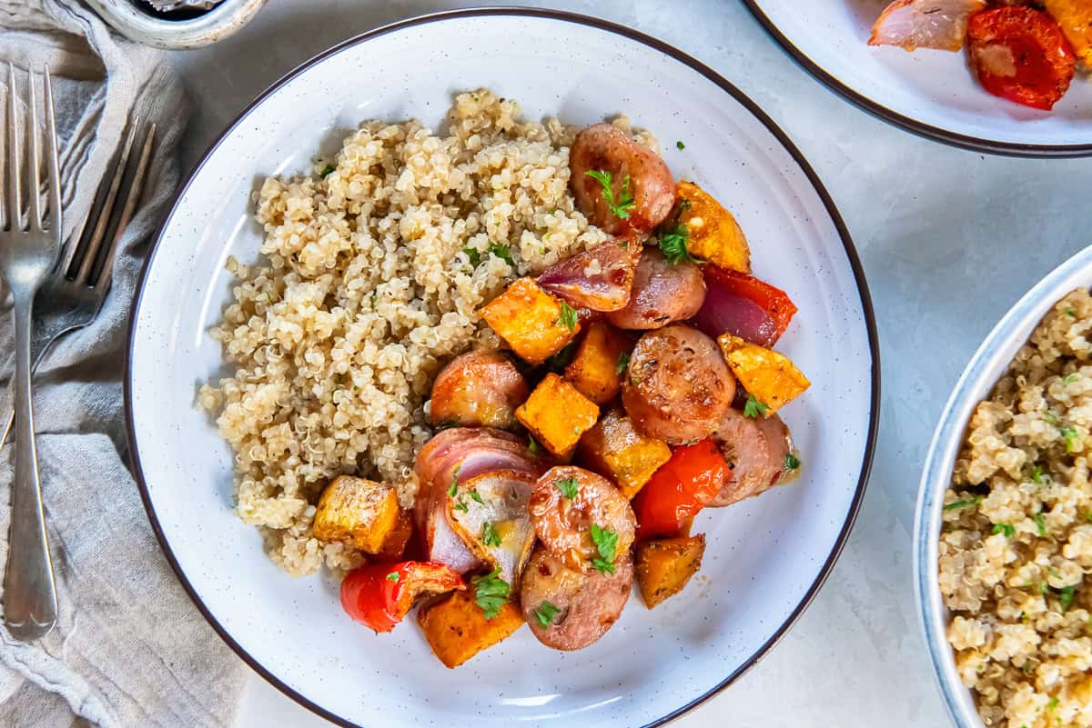 Roasted chicken sausage, sweet potatoes, red bell peppers, and red onion served alongside fluffy quinoa on a white plate, with additional quinoa and a second plate partially visible.