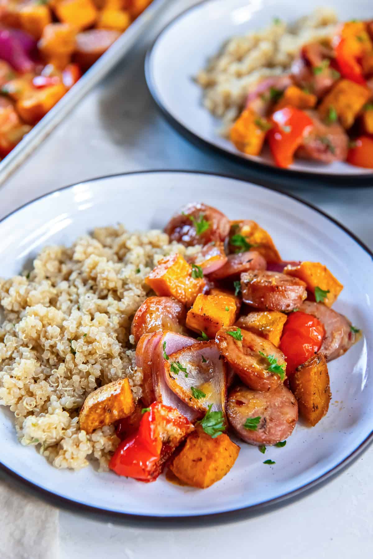 Sliced chicken sausage, roasted sweet potatoes, red bell peppers, and red onion served with fluffy quinoa on a white plate, with a second plate in the background.