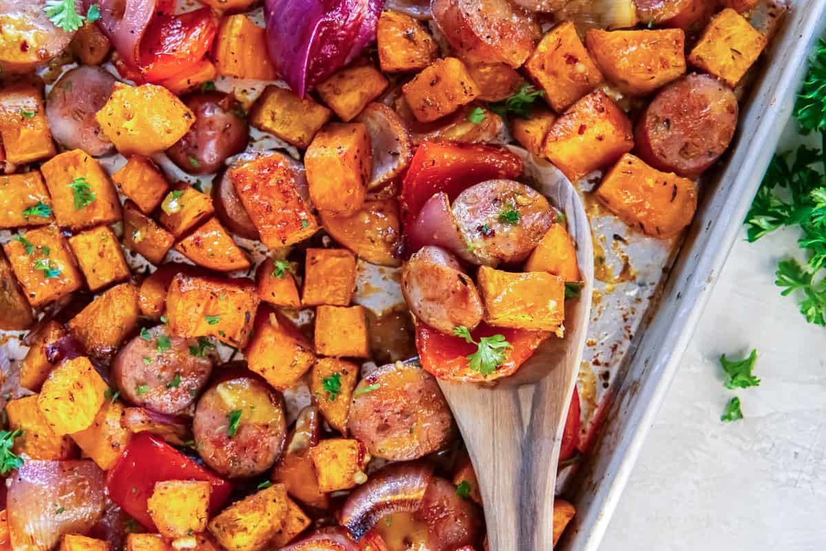 Sheet pan sausage and sweet potatoes with roasted red bell peppers and red onion, with a wooden spoon holding a serving of the colorful mixture.