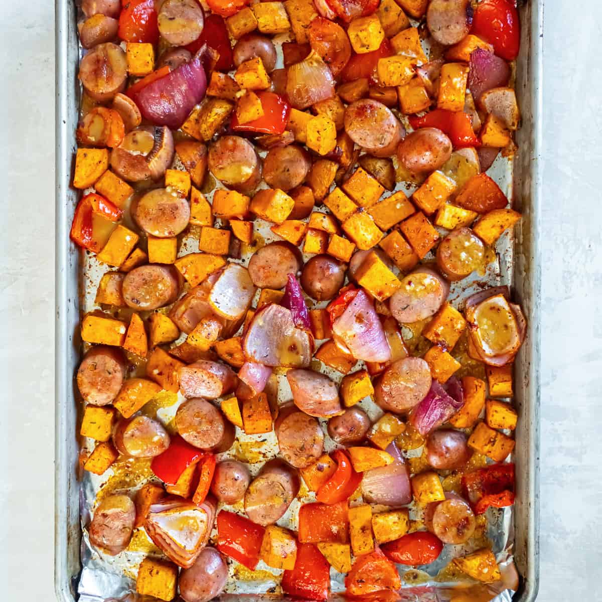 Roasted chicken sausage, sweet potatoes, red bell pepper, and red onion coated in a glossy maple-mustard glaze on a baking sheet.