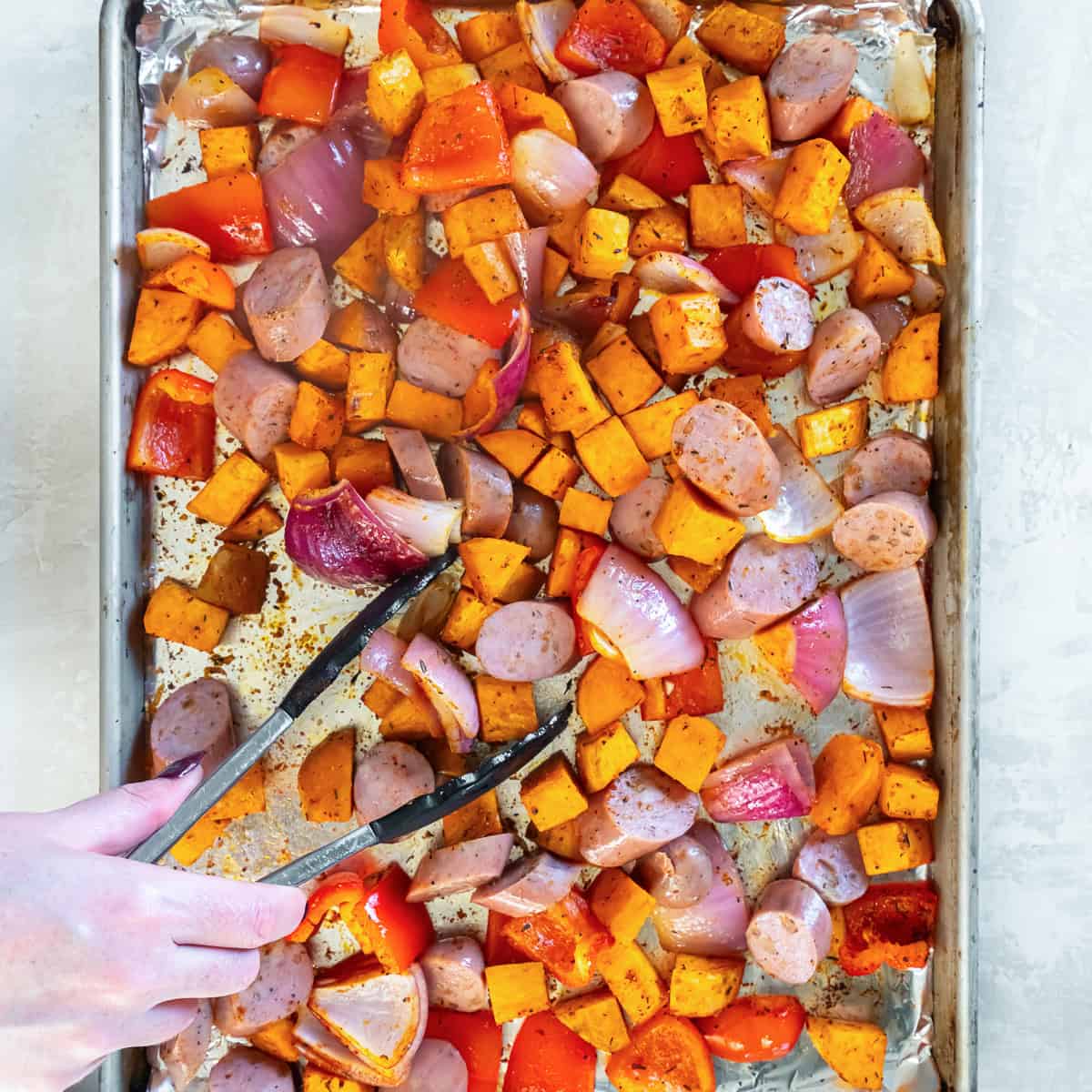 Sliced chicken sausage, cubed sweet potatoes, red bell pepper, and red onion being tossed with tongs on a foil-lined baking sheet.
