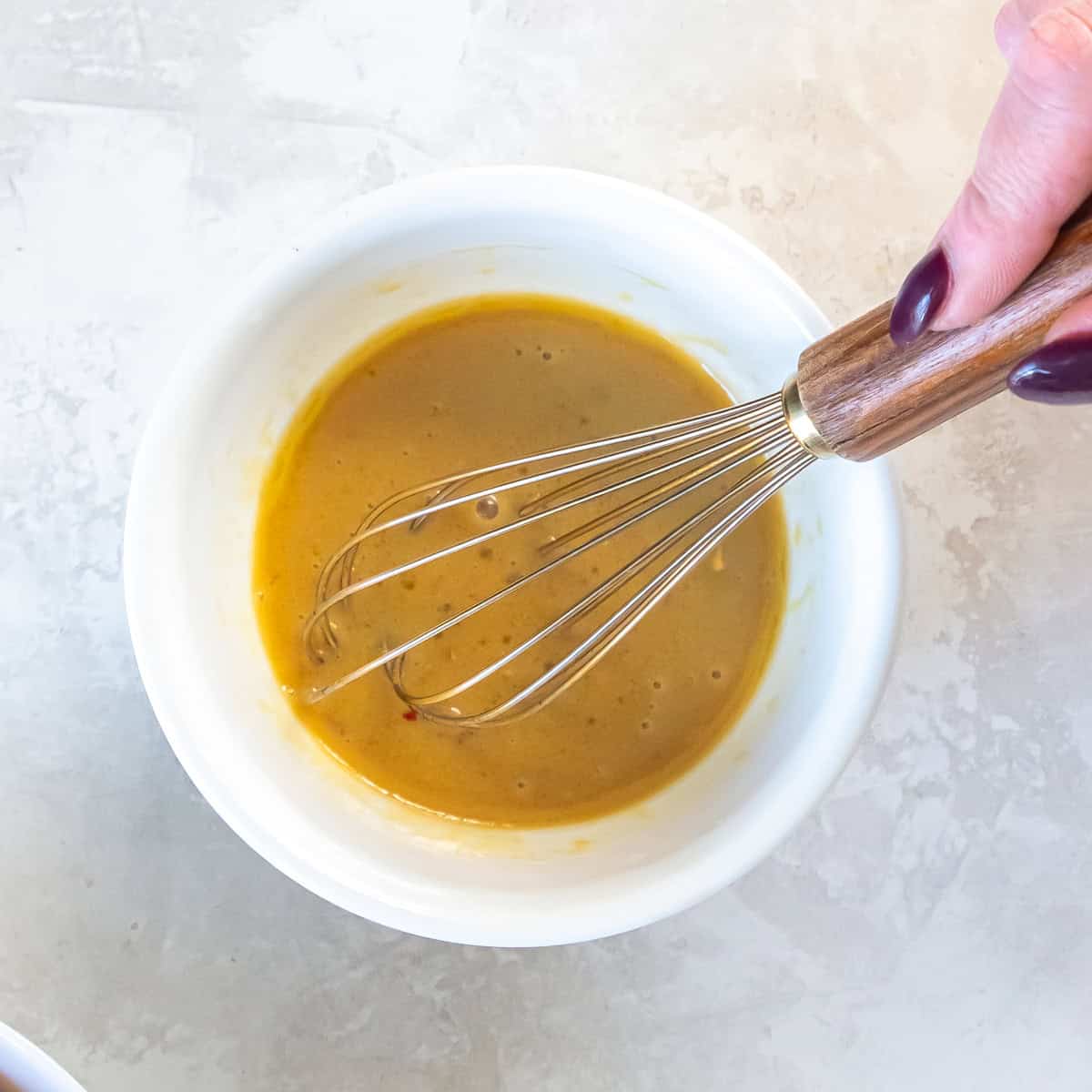 Golden maple-mustard glaze being whisked in a white bowl before adding it to the sheet pan chicken sausage and sweet potatoes.