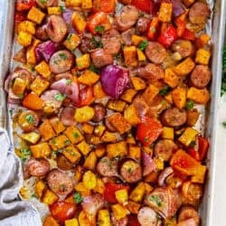 A foil-lined baking sheet filled with sliced chicken sausage, roasted sweet potatoes, red bell peppers, and red onions, finished with a maple mustard glaze and chopped parsley.