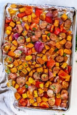 A foil-lined baking sheet filled with sliced chicken sausage, roasted sweet potatoes, red bell peppers, and red onions, finished with a maple mustard glaze and chopped parsley.